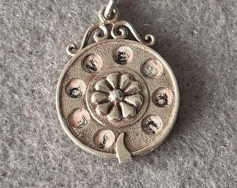 May include: A silver-toned pendant with a rotary phone design. The pendant features a central flower design and a series of small circles around the perimeter, each with a number or symbol. The pendant has a loop for attaching to a chain.
