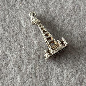 May include: A silver-toned metal charm shaped like the Eiffel Tower. The charm features an openwork lattice design throughout the structure, a pointed top, and a small circular jump ring attached at the apex for hanging on a chain or bracelet.