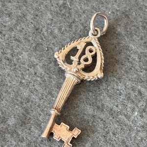 Könnte beinhalten: Silberner Schlüsselanhänger mit der Zahl 18 in der Mitte des Schlüsselloches. Der Schlüssel ist traditionell gestaltet mit einem dekorativen Kopf und einem einfachen, geraden Schaft.