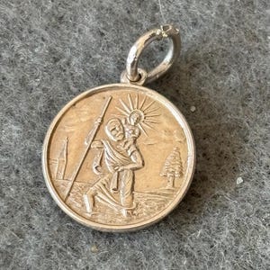 Op de afbeelding: Een zilveren hanger met een gedetailleerd reliëf van Sint-Christoffel die het Christuskind draagt. De ronde hanger heeft een lus voor een ketting en toont een staf, een boom en een kerk. De hanger ligt op een grijze ondergrond.