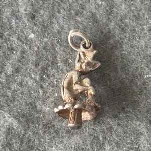 May include: A silver-toned charm depicting a seated gnome figure on a mushroom. The gnome is wearing a hat and has its legs crossed. The charm has a loop at the top for attachment to a bracelet or necklace.