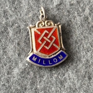 May include: Silver charm featuring a red and white enamel design of a knot within a shield shape. The charm has a blue enamel band around the bottom with the word "MILLOM" in white lettering.