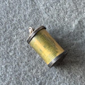 Peut inclure: Pendentif cylindrique avec un dessus et un dessous en argent. Le corps du pendentif est doré avec un motif de soleil vert et jaune. Le mot "SILVER" est estampé sur le bord supérieur.
