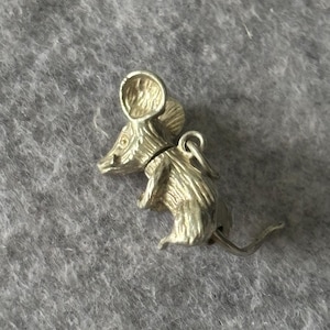 Może przedstawiać: Srebrny charms w kształcie myszy z detalami, w tym dużymi uszami, spiczastym nosem i zakręconym ogonem. Charms ma małą pętlę do przymocowania do bransoletki lub naszyjnika. Mysz jest przedstawiona w pozycji siedzącej.