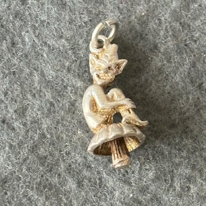 Puede incluir: Encanto de plata de un duende sentado en un hongo. El duende tiene orejas puntiagudas y una expresión traviesa.