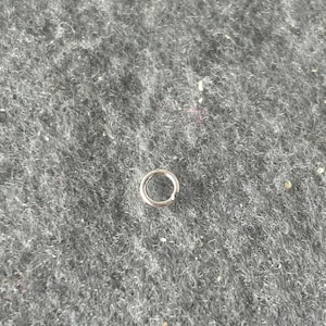 May include: A small, silver-colored metal jump ring. The ring is circular and has a smooth, shiny surface. It is lying on a textured, gray surface.