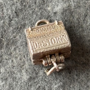 Pode incluir: Um berloque de prata em forma de mala pequena. O berloque tem as palavras "DR STORK" gravadas na frente. A mala é articulada e tem um pequeno laço na parte superior para prender a uma pulseira ou colar.