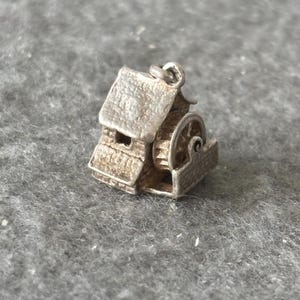 May include: A small, silver-toned charm in the shape of a water mill. The charm features a detailed roof, walls, and a water wheel. The charm is likely made of metal and has a loop for attaching to a bracelet or necklace.