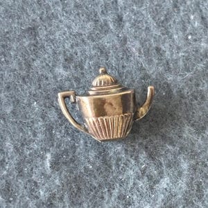 K&ouml;nnte beinhalten: Ein kleiner, goldfarbener Teekannen-Anstecker mit detailliertem Design. Der Anstecker hat einen Griff, einen Ausguss und einen Deckel mit einem kleinen Knopf. Der K&ouml;rper der Teekanne hat vertikale Linien. Der Anstecker befindet sich vor einem grauen, strukturierten Hintergrund.
