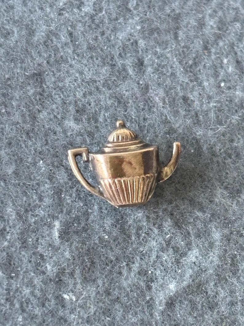 K&ouml;nnte beinhalten: Ein kleiner, goldfarbener Teekannen-Anstecker mit detailliertem Design. Der Anstecker hat einen Griff, einen Ausguss und einen Deckel mit einem kleinen Knopf. Der K&ouml;rper der Teekanne hat vertikale Linien. Der Anstecker befindet sich vor einem grauen, strukturierten Hintergrund.
