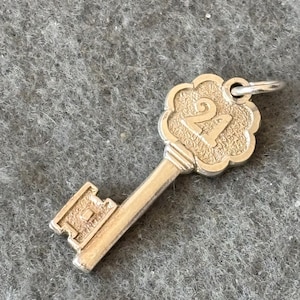 Op de afbeelding: Een zilveren sleutelbedel met een decoratief hoofd met het cijfer "2" in een hartvormig ontwerp. De sleutel is een populaire bedel voor het maken van sieraden.