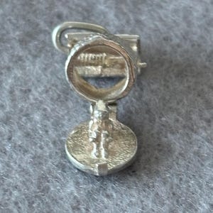 May include: A small, silver-colored charm with a detailed design. The charm features a circular element with an engraved pattern, connected to a rounded base with a figure. A loop is at the top for attachment.