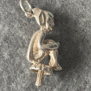 May include: Silver charm of a small elf sitting on a mushroom. The elf has pointed ears and is holding its hands together. The charm has a loop at the top for attaching to a bracelet or necklace.