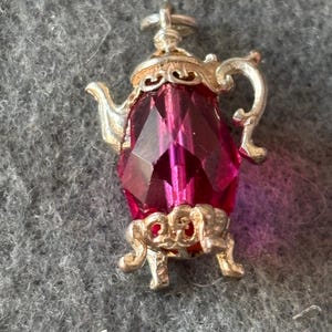 Könnte beinhalten: Ein silberfarbener Anhänger in Form einer Teekanne mit einem facettierten, tiefrosa Edelsteinkörper. Die Teekanne hat verzierte Details an Griff, Ausguss und Boden, mit einer kleinen Schlaufe oben zur Befestigung. Der Anhänger ist vor grauem Hintergrund platziert.
