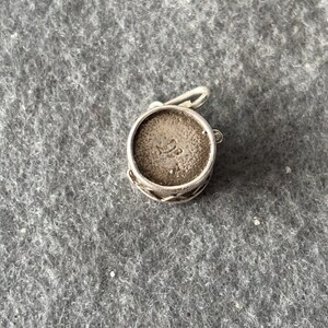 May include: A small, round, silver-toned charm with a decorative filigree band. The charm has a loop for attaching to a bracelet or necklace. The interior of the charm has a textured surface with an embossed design.