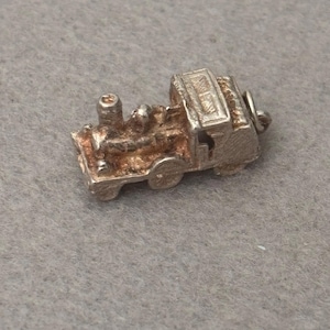 May include: A small, three-dimensional silver-toned metal charm shaped like a steam locomotive train. The charm features a textured surface, a prominent smokestack, a cab section, and a small circular jump ring attached to the rear for hanging.