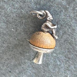 May include: A silver charm featuring a small figure sitting on a mushroom. The figure is wearing a pointed hat and has a long, flowing beard. The mushroom is made of wood and has a brown color.