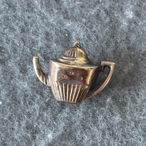 K&ouml;nnte beinhalten: Ein kleiner, goldfarbener Teekannen-Anh&auml;nger. Der Anh&auml;nger hat einen Henkel, einen Ausguss und einen Deckel. Der Boden der Teekanne hat vertikale Linien. Der Anh&auml;nger ist wahrscheinlich aus Metall und vor einem grauen Hintergrund platziert.