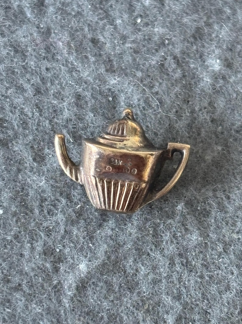 K&ouml;nnte beinhalten: Ein kleiner, goldfarbener Teekannen-Anh&auml;nger. Der Anh&auml;nger hat einen Henkel, einen Ausguss und einen Deckel. Der Boden der Teekanne hat vertikale Linien. Der Anh&auml;nger ist wahrscheinlich aus Metall und vor einem grauen Hintergrund platziert.