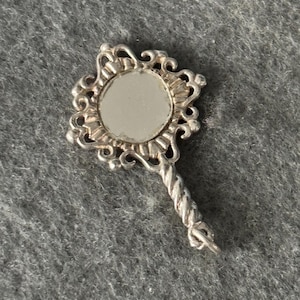 May include: A small, ornate silver-colored hand mirror. The mirror has a round reflective surface surrounded by a decorative, swirling frame. The handle is twisted and has a small loop at the end.
