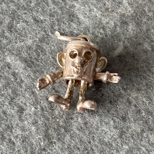 May include: A silver-toned charm in the shape of a cartoon character with a cylindrical body, large eyes, and outstretched arms. The charm has a small hat and detailed facial features, including a smiling mouth and ears. The charm is on a gray, textured surface.