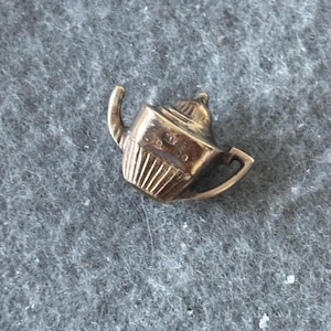 K&ouml;nnte beinhalten: Ein kleiner, goldfarbener Teekannen-Anh&auml;nger. Der Anh&auml;nger hat einen gebogenen Ausguss, einen Henkel und einen Deckel mit einem kleinen Knopf. Der K&ouml;rper der Teekanne hat vertikale Linien. Der Anh&auml;nger ist wahrscheinlich aus Metall.