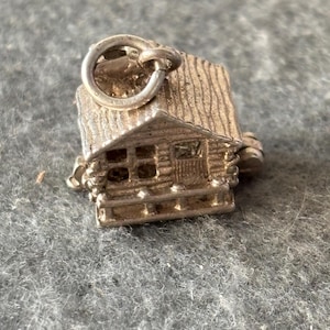 May include: A silver-colored charm in the shape of a small cabin. The cabin has a textured roof, a small porch, and windows. The charm is attached to a silver ring.
