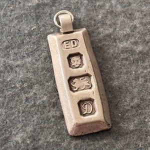 May include: A silver-coloured metal pendant shaped like a small rectangular ingot. The front surface features four distinct stamped marks: the letters ED, a leopard head, a walking lion, and the letter D, all enclosed within individual recessed squares.
