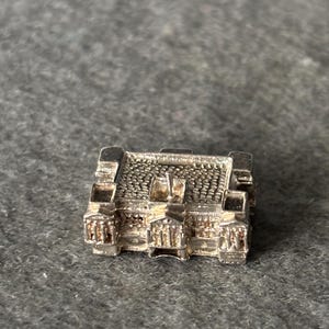 May include: A small, detailed silver-colored charm in the shape of a building, possibly a miniature replica of a structure. The charm has intricate architectural details, including a textured roof and facade.