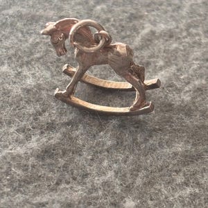 May include: A rose gold-toned metal charm shaped like a rocking horse. The charm features a detailed horse figure with a mane and tail, mounted on two curved rockers. A small circular jump ring is attached to the top of the horse's back for hanging.