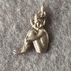 May include: A silver-toned metal charm shaped like a seated figure with pointed ears and a cap, depicted in a curled position with arms wrapped around the knees. The charm includes a small circular jump ring at the top for attachment to a chain.