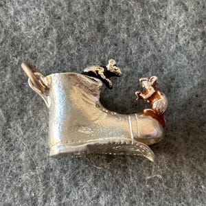 May include: A silver-colored boot charm with two small, detailed mice. One mouse is black and the other is brown. The boot has a chain attached to the top. The charm is likely made of metal.