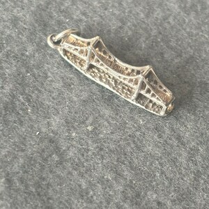 May include: A silver charm shaped like a bridge. The charm is detailed and has a loop at the top for attaching to a bracelet or necklace.
