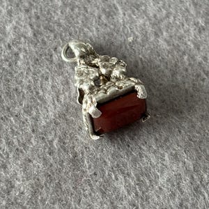 May include: A silver pendant with a rectangular, reddish-brown stone. The pendant has ornate detailing around the stone and a small loop at the top for a chain. The pendant is likely vintage or antique.