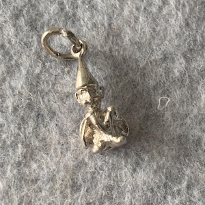Op de afbeelding: Een kleine, zilverkleurige bedel in de vorm van een kabouter of elf. De bedel heeft een puntmuts en houdt een voorwerp vast. Hij is bevestigd aan een kleine ring om op te hangen.