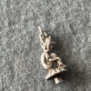 May include: A silver charm featuring a fairy sitting on a mushroom. The fairy has a pointed hat and is holding a small object in her hands.