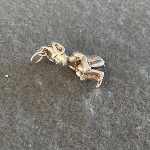Puede incluir: Un charm de color plateado con forma de figura sentada, posiblemente una criatura mitológica. El charm tiene un lazo para sujetarlo a una pulsera o collar. La figura está detallada con rasgos faciales y extremidades visibles.