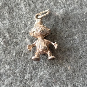 Op de afbeelding: Een kleine, zilverkleurige bedel in de vorm van een grillige kabouter of elf. De bedel heeft een puntmuts, gedetailleerde gezichtskenmerken en uitgestrekte armen. Het heeft een lus aan de bovenkant om aan een ketting of armband te bevestigen. De bedel ligt op een grijze ondergrond.