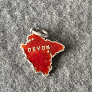 May include: A silver-toned charm in the shape of Devon, England, with a bright red enamel fill. The word "DEVON" is written in white letters across the charm. The charm has a small loop for attaching to a bracelet or necklace.