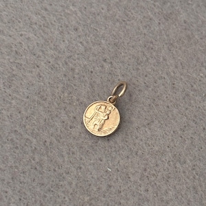 May include: A small, round, gold-toned metal pendant featuring an embossed design of a figure carrying a staff and a smaller figure on their shoulder. The pendant includes a circular jump ring at the top for attachment to a chain or cord.