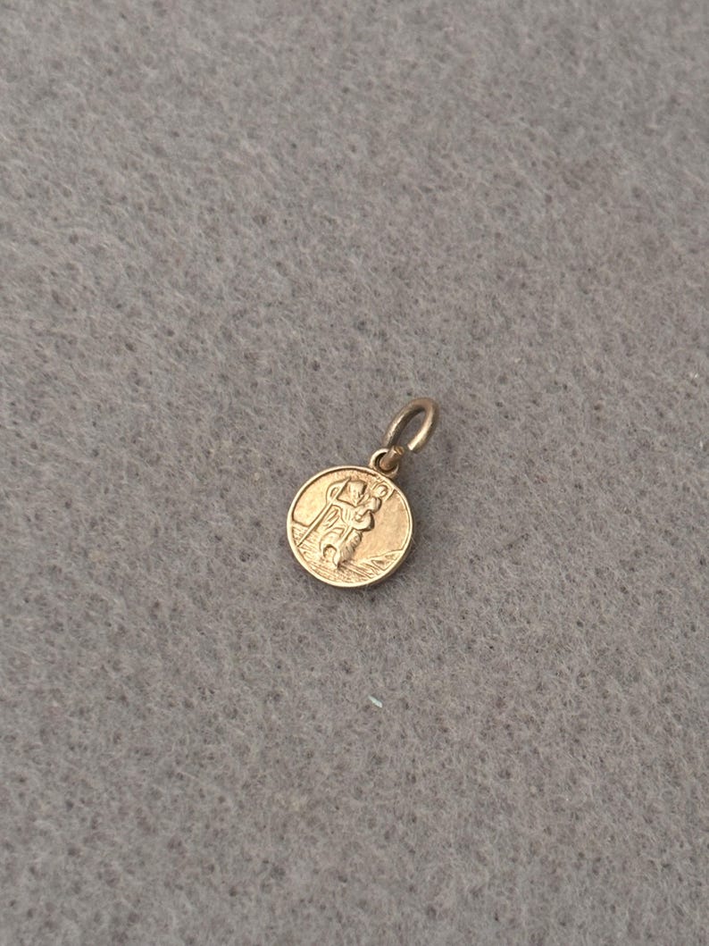 May include: A small, round, gold-toned metal pendant featuring an embossed design of a figure carrying a staff and a smaller figure on their shoulder. The pendant includes a circular jump ring at the top for attachment to a chain or cord.