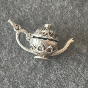 Könnte beinhalten: Ein silberner Anhänger in Form einer Teekanne mit einem verzierten Design. Die Teekanne hat einen Klappdeckel und einen Ausguss. Der Anhänger ist für ein Armband oder eine Halskette geeignet.
