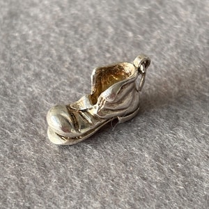 Op de afbeelding: Een kleine, zilverkleurige laarshanger met ingewikkelde details. De hanger heeft een afgeronde neus, een getextureerd oppervlak en een lus om aan een armband of ketting te bevestigen. De laars is een miniatuurweergave van een werkschoen.