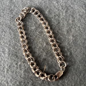 May include: Silver-coloured double-link chain bracelet with a lobster clasp. The bracelet is made of small, interconnected oval links. The jewellery is displayed on a grey surface.