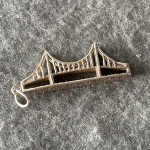 May include: A silver bridge charm with intricate detailing, including support cables and the text "SEVERN ROAD BRIDGE". The charm is designed with a loop for attaching to a bracelet or necklace. The bridge design is a classic style.
