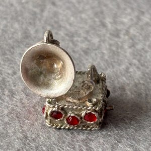 Op de afbeelding: Een kleine, zilverkleurige bedel in de vorm van een vintage grammofoon. De bedel heeft een uitlopende hoorn, een vierkante basis met rode edelsteenaccenten en een lus om te bevestigen. De grammofoonbedel is een miniatuurreplica van een klassieke muziekspeler.
