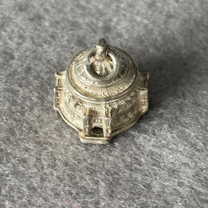 Może przedstawiać: Srebrzysty metalowy charms w kształcie miniaturowego budynku z kopułą, posiadający detale architektoniczne, takie jak łukowate okna i kolumny. Charms posiada okrągłe kółeczko na górze, umożliwiające zawieszenie go na łańcuszku lub bransoletce.