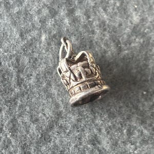 May include: A silver charm in the shape of a crown. The crown has a detailed design and a loop at the top for attaching to a bracelet or necklace.