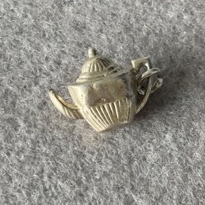 Könnte beinhalten: Ein kleiner, silberfarbener Teekannen-Anhänger mit detailliertem Design. Der Anhänger hat einen Ausguss, einen Henkel und einen Deckel und ist wahrscheinlich aus Metall gefertigt. Die Teekanne hat eine klassische Form und eignet sich für die Schmuckherstellung oder als Anhänger.