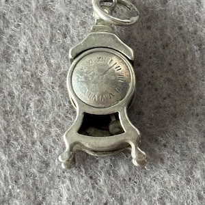 May include: A silver-toned charm in the shape of a miniature clock. The clock face features Roman numerals and hands. The charm has a small loop at the top for attachment and rests on four small feet.